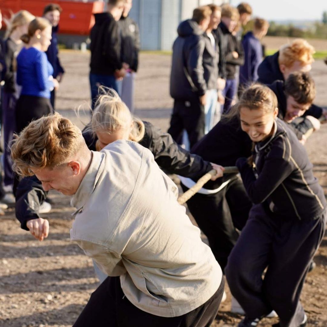 En gruppe efterskoleelever deltager i tovtrækning udendørs. Eleverne trækker i et reb med stor energi, mens andre elever står omkring og følger med.